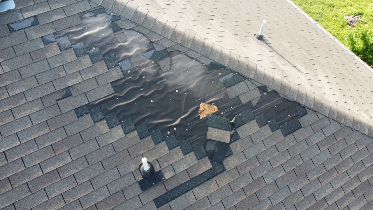 Drone close-up of wind-damaged shingles torn off a roof, exposing the underlayment